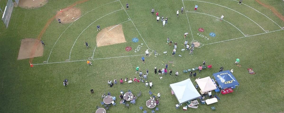 An image showing an aerial shot of a wiffle ball field. participants in a tournament playing the game. There is also shade tents and table set up for spectators