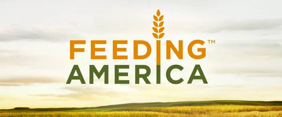 Logo for feeding America. Text is in orange and green colors. The I in feeding is wheat stock. The background image of an open field