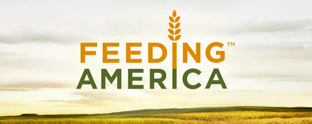 Logo for feeding America. Text is in orange and green colors. The I in feeding is wheat stock. The background image of an open field