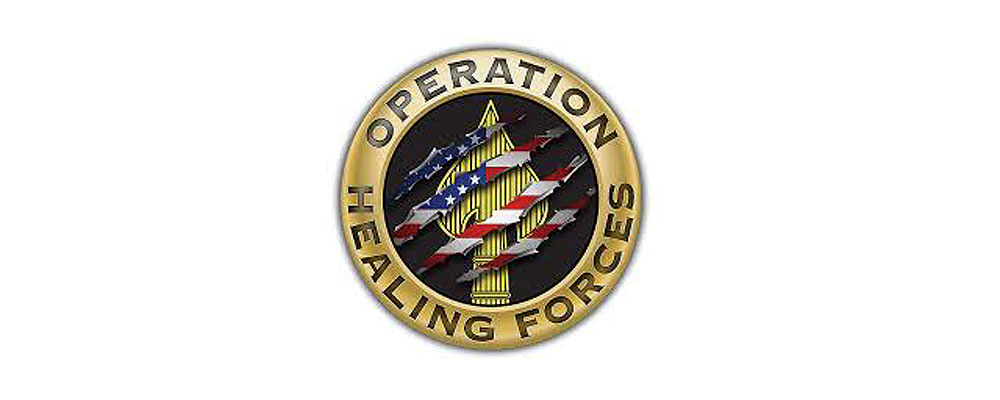 Logo for Operation Healing Forces. Logo appears in a badge style with a circle design. Text wraps around the metal rim. In the center is a yellow spade design. Tear marks are shown over top of the image showing an American flag underneath.