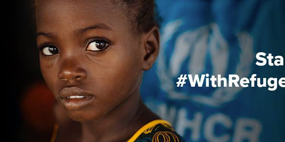 Close up Image of young refugee girl face looking into the camera. The background is blurred out. The words “Stand” and “#WithRefugees” are written in white.