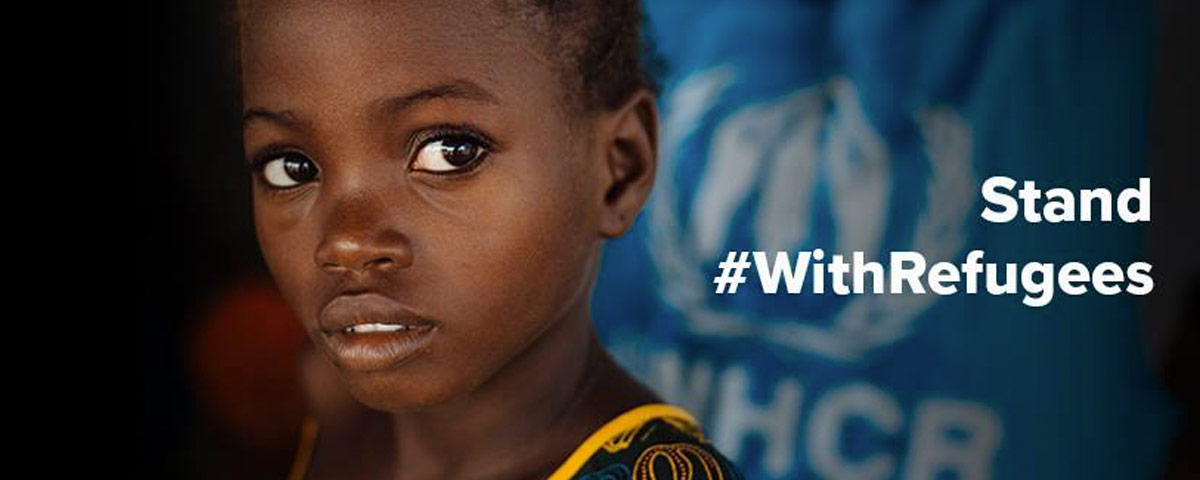 Close up Image of young refugee girl face looking into the camera. The background is blurred out. The words “Stand” and “#WithRefugees” are written in white.